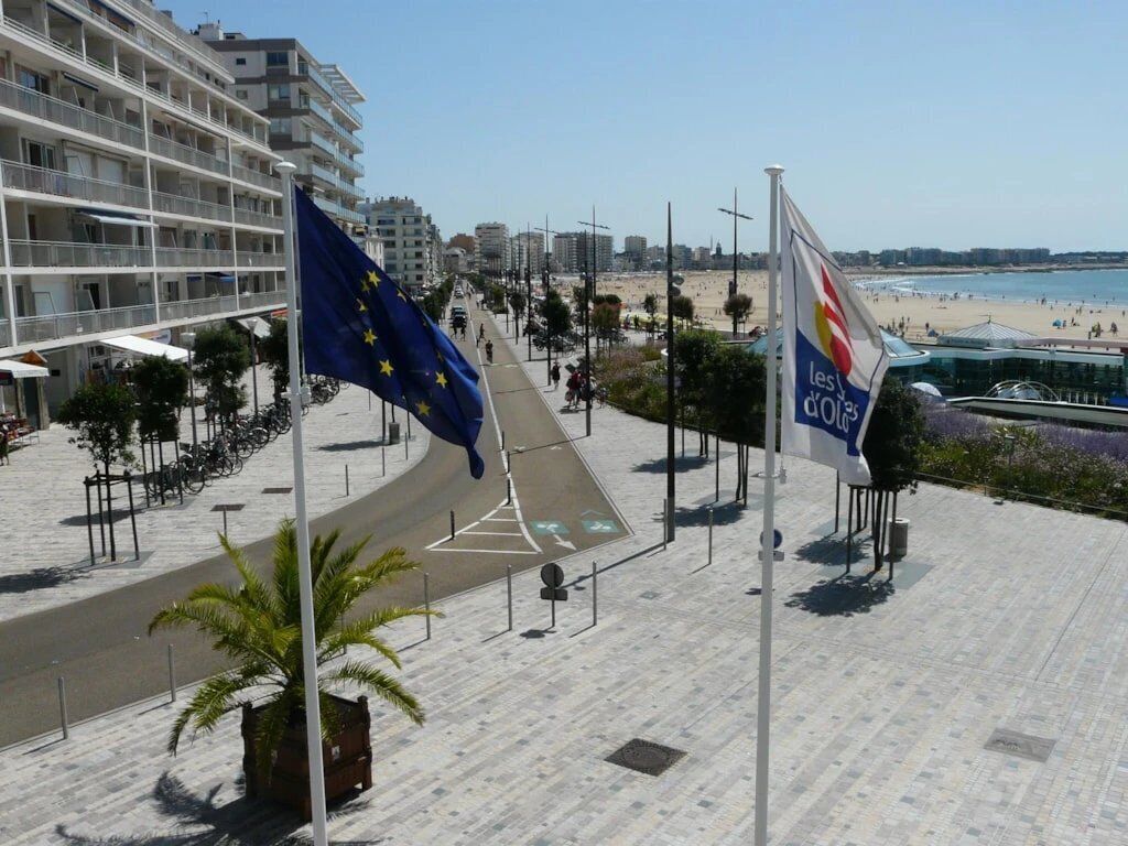 Un passeig amb banderes, una platja i edificis al fons.