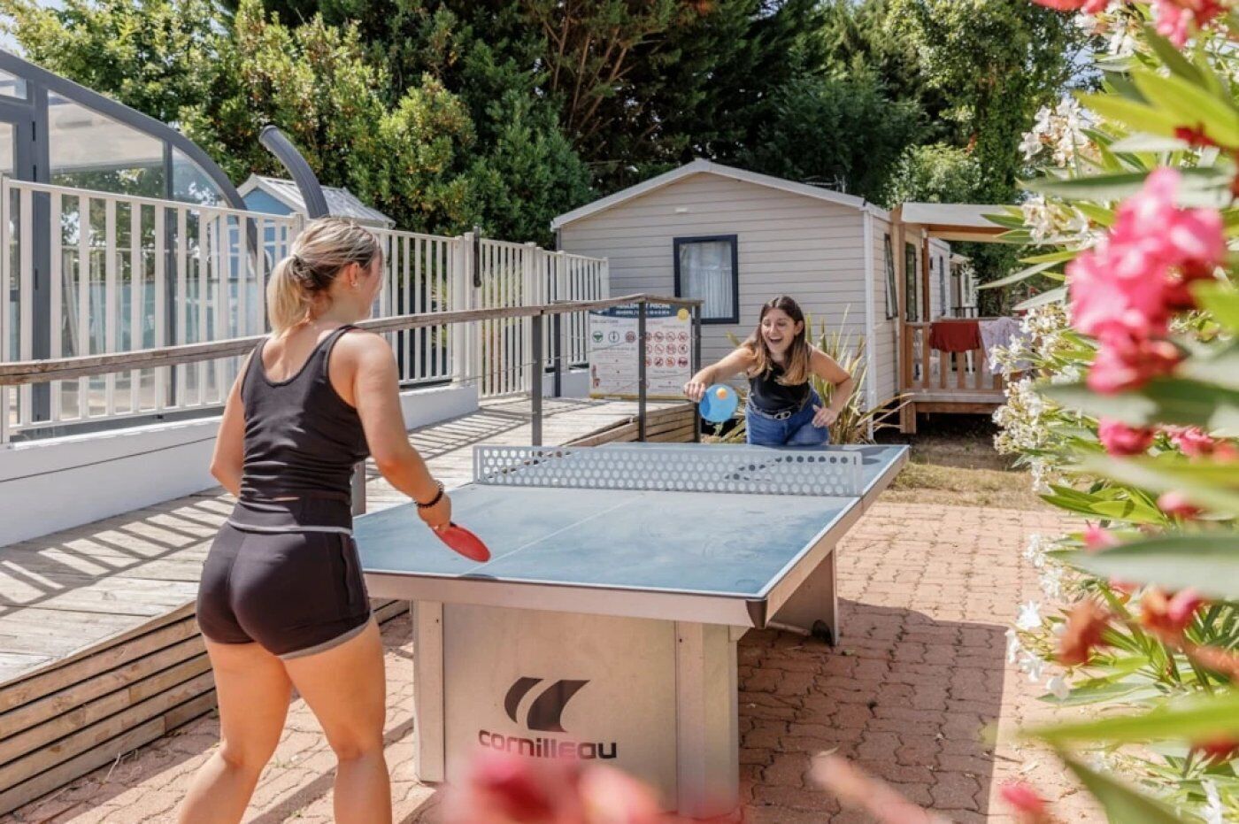 Twee personen spelen tafeltennis buiten, met een huis op de achtergrond.