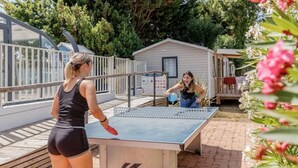 Zwei Personen spielen Tischtennis draußen, mit einem Haus im Hintergrund.