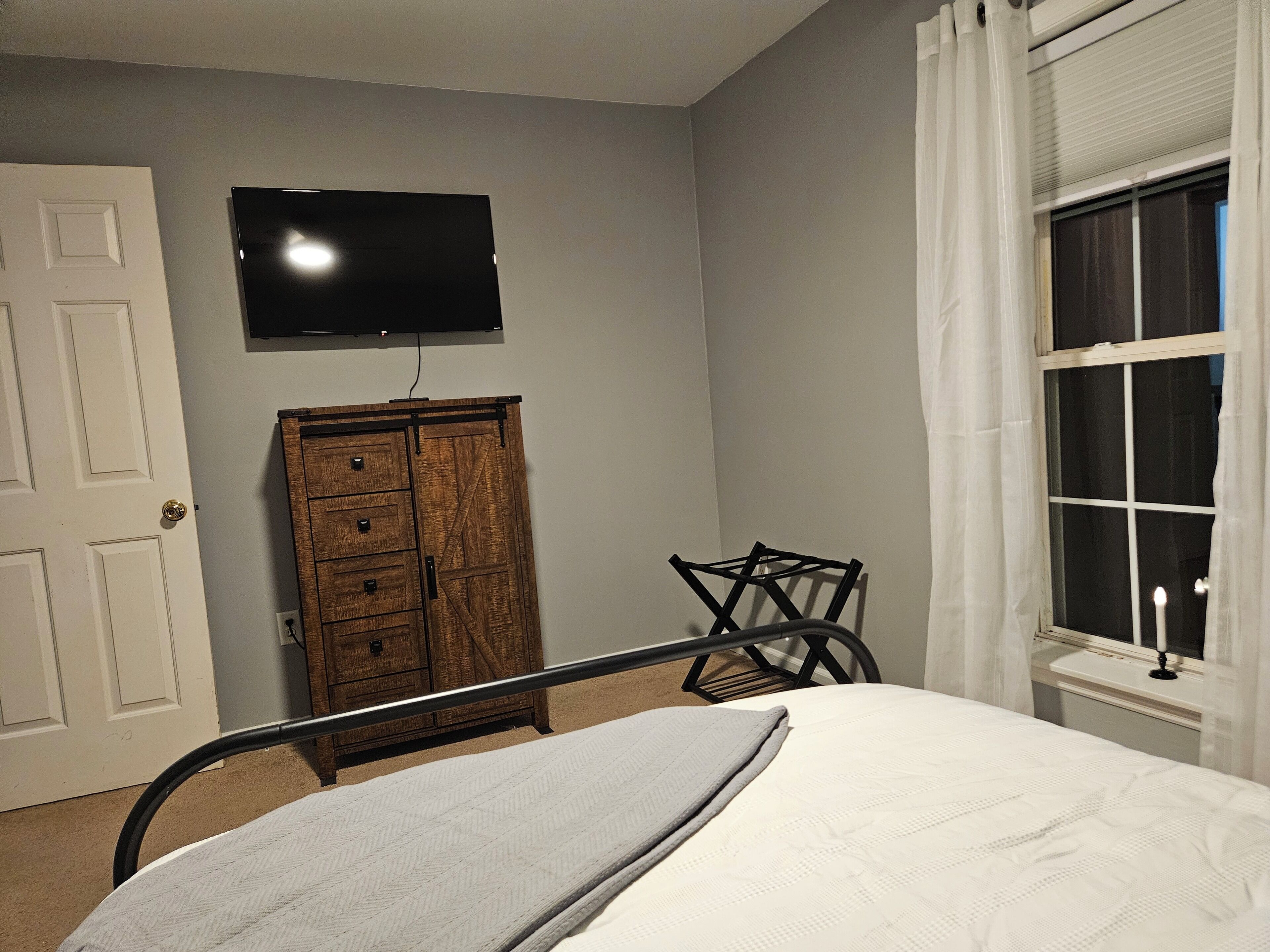 Ein Schlafzimmer mit einem Bett, einer Holzkommode, einem Sessel, einem an der Wand befestigten Fernseher und einem Fenster mit Vorhängen.