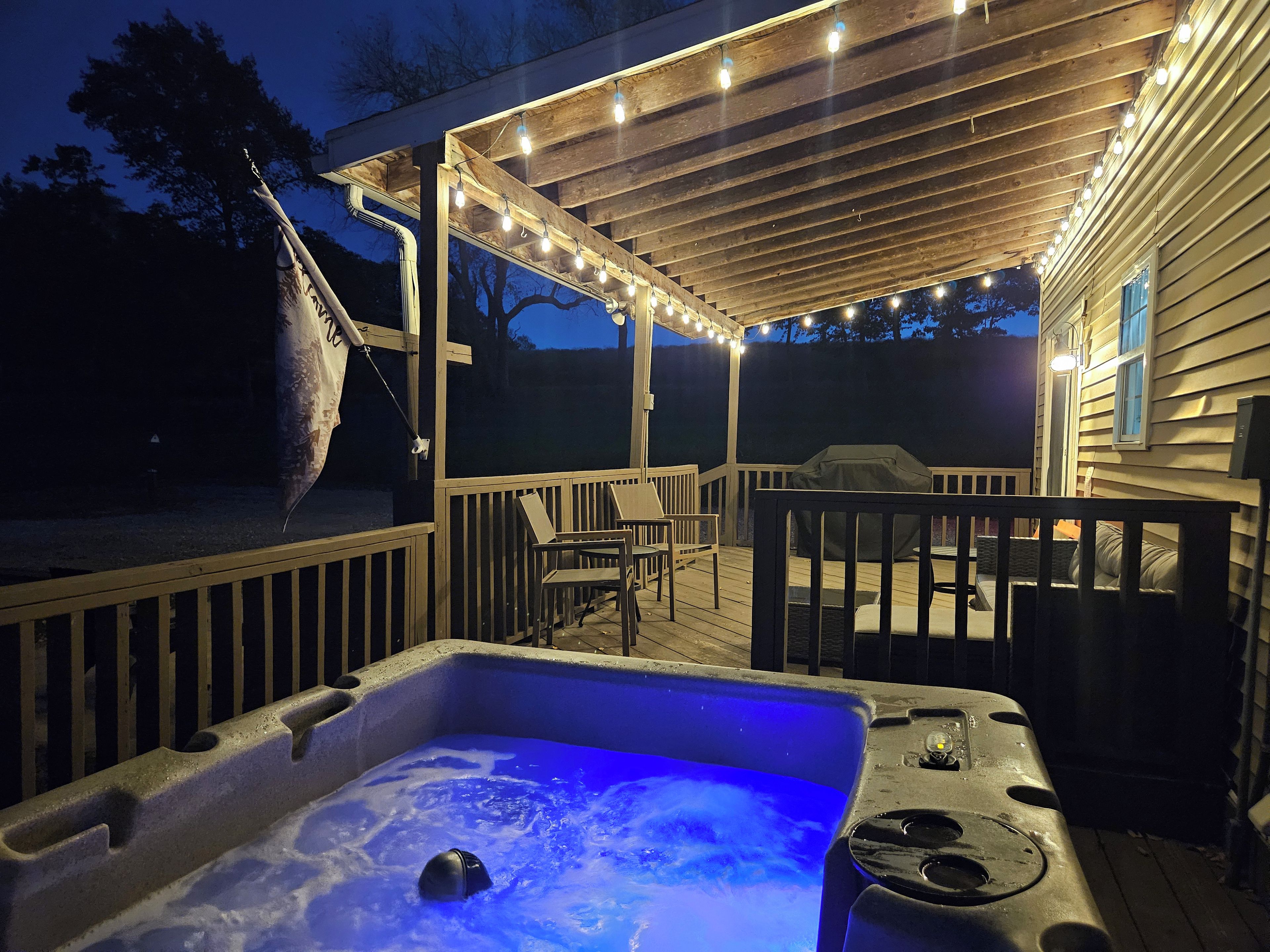 A hot tub with a blue light, a wooden deck, outdoor furniture, and string lights.