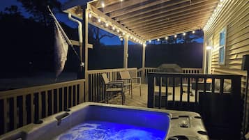A hot tub with a blue light, a wooden deck, outdoor furniture, and string lights.