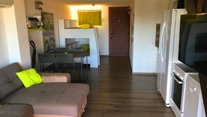 A compact living space with a sofa, dining table, chairs, a kitchen area with a refrigerator, and a television. - Magnifique Appartement  VUE MER et MONTAGNES (Ghisonaccia)