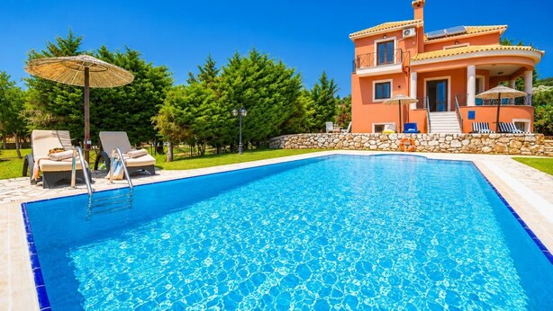 Ein Swimmingpool mit Liegestühlen und Sonnenschirmen, eine Steinmauer und ein zweistöckiges Gebäude mit Balkon.