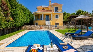 A swimming pool with lounge chairs, a stone wall, and a house with a balcony in the background. - Ionian Blue - Three Bedroom Villa, Sleeps 7 (Svoronata)