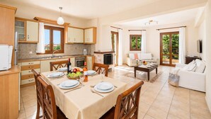 A spacious living area with a dining table set for four, a kitchen with wooden cabinets, a TV, and a balcony with a view of greenery. - Ionian Blue - Three Bedroom Villa, Sleeps 7 (Svoronata)