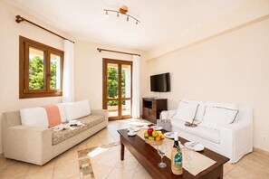 A living room with two sofas, a coffee table with fruit and bottles, a TV, and a sliding glass door leading to a balcony.