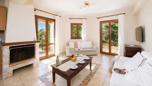 A spacious living area with a fireplace, a sofa, a dining table set with plates and a bottle, and a TV on a wooden stand. - Ionian Blue - Three Bedroom Villa, Sleeps 7 (Svoronata)
