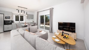Ein modernes Apartment mit Wohnzimmer, Essbereich und Küche. Ein großes Fenster mit Vorhängen, ein Flachbildfernseher und ein Sofa sind vorhanden.