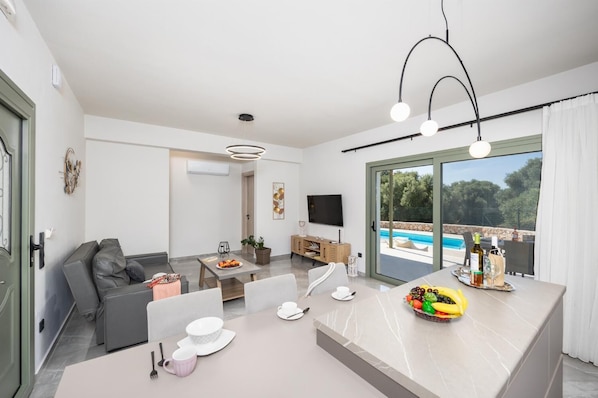Ein modernes Wohnzimmer mit einem grauen Sofa, einem Esstisch mit Obstschale und einem Fernseher.
