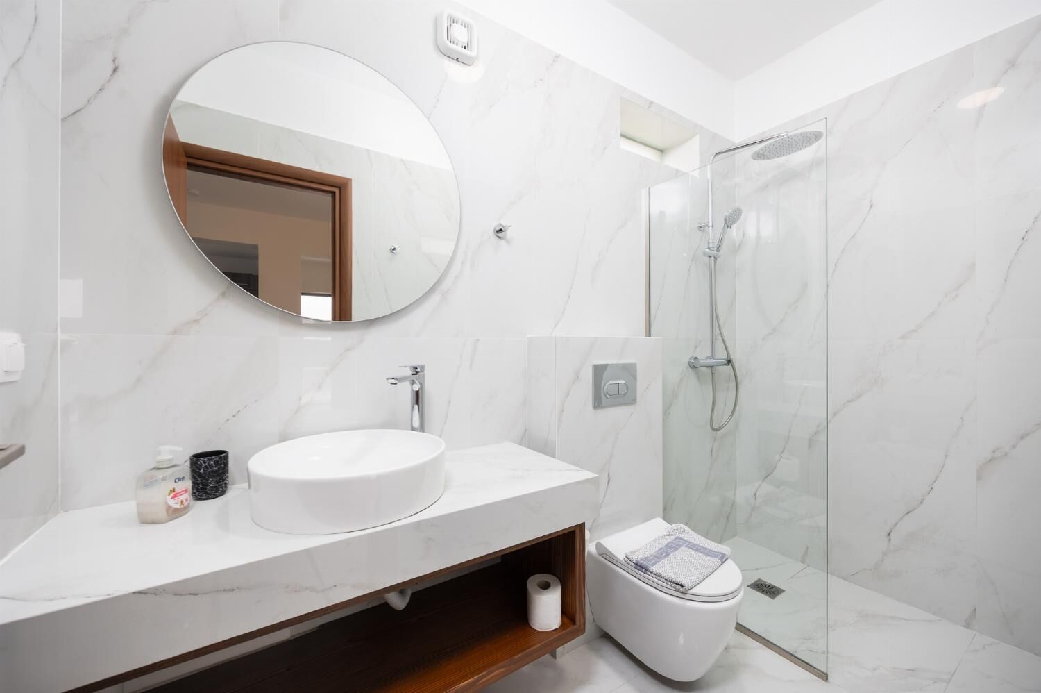 Een moderne badkamer met een ronde spiegel, witte marmeren wanden, een witte wastafel en een douchecabine met glazen wanden.