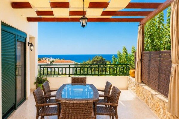 Eine überdachte Terrasse mit Tisch und Stühlen und Blick auf das Meer, sowie eine Holz-Pergola.