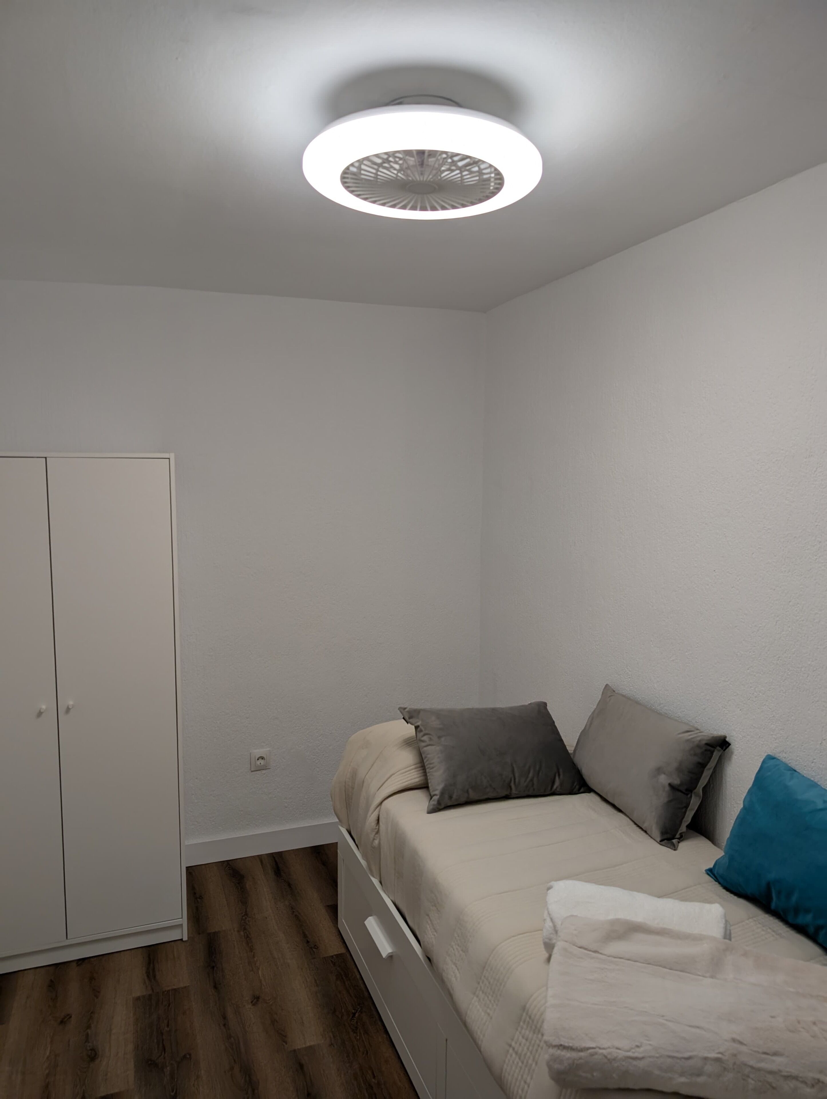 A small, single-bed room with a wooden floor, a white wardrobe, and a ceiling light fixture.