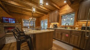 A cozy kitchen with wooden cabinetry, a large island with bar stools, a stone fireplace, and a stone wall-mounted TV. - 3 Bed | 4 Bath Mountain Escape in Wears Valley (Sevierville)