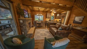 A cozy cabin interior with a stone fireplace, wooden ceiling beams, a dining area, and a living room with a leather sofa and green armchairs. - 3 Bed | 4 Bath Mountain Escape in Wears Valley (Sevierville)