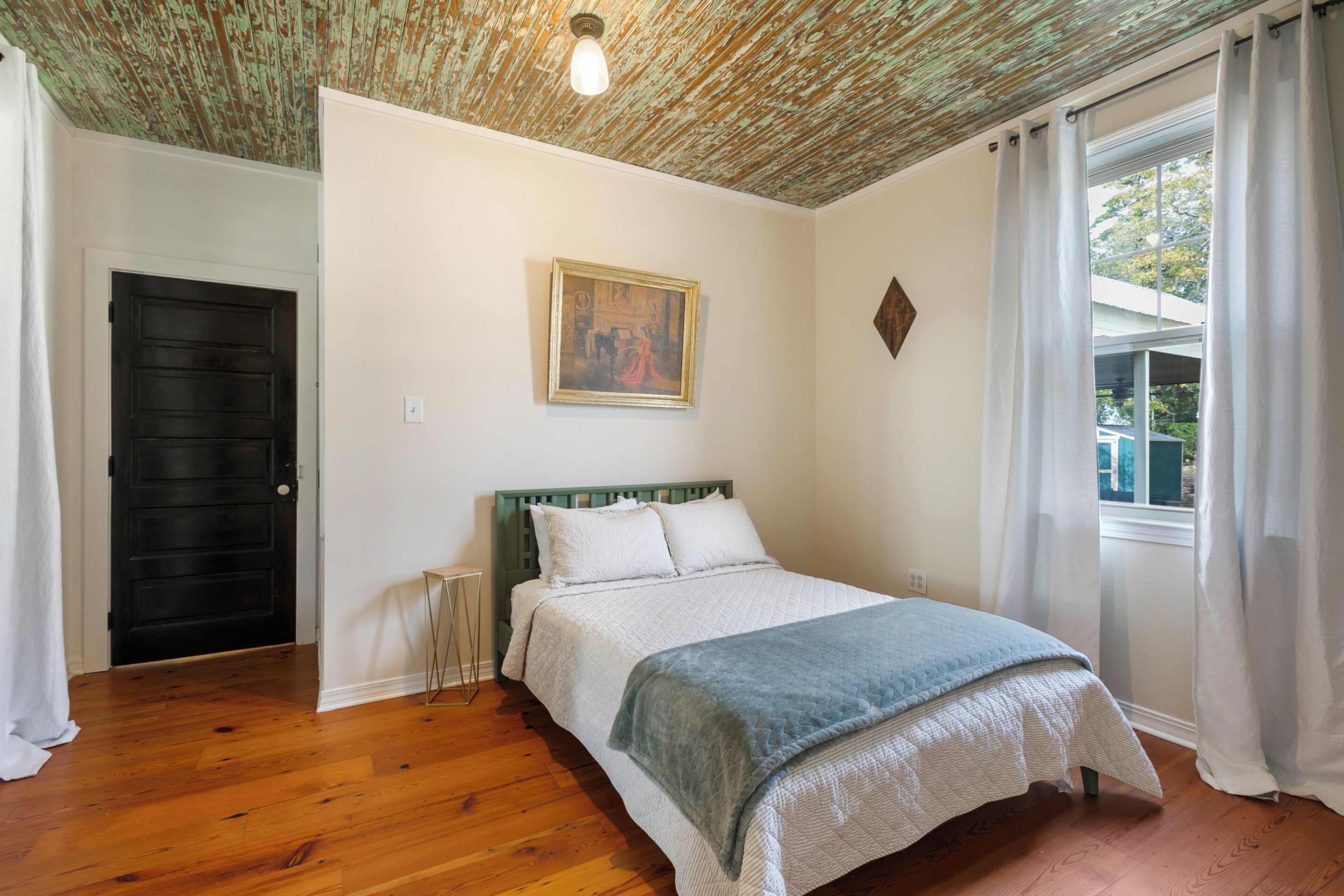 A bedroom with a wooden floor, a bed with white bedding, a green headboard, a painting on the wall, and a window with curtains.