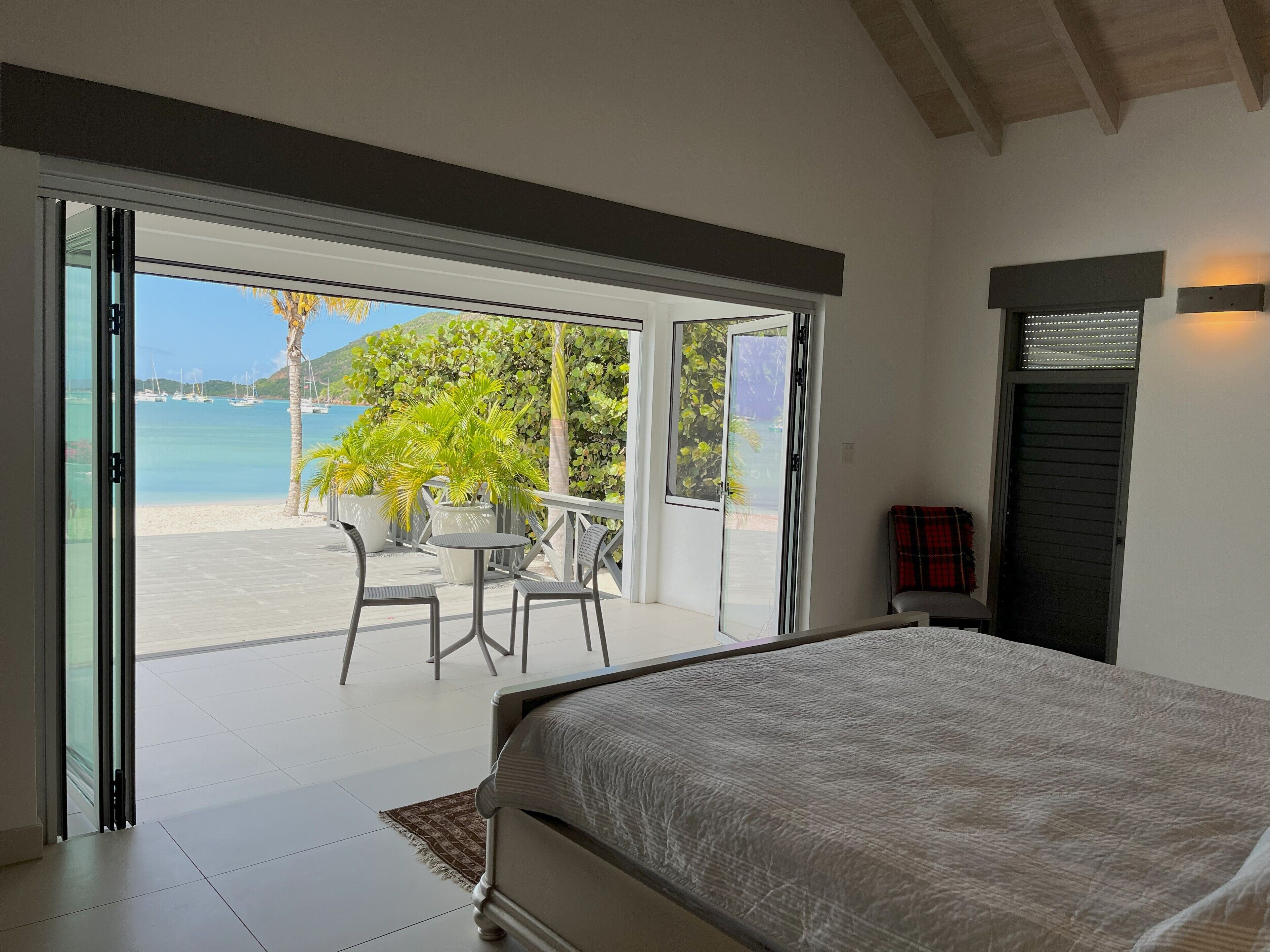 Un dormitorio con una cama grande, un ventanal corredero que da a un balcón con mesa y sillas, y vista a la playa y barcos.