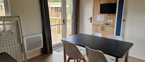 A compact room with a dining table, two chairs, a TV mounted on the wall, and a sliding glass door leading to an outdoor area.