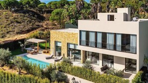 A modern two-story house with a swimming pool, outdoor seating, and a view of a hilly landscape.