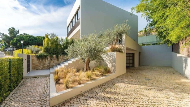 A modern building with a flat roof, a stone pathway leading to the entrance, and a small garden area with a tree and shrubs.
