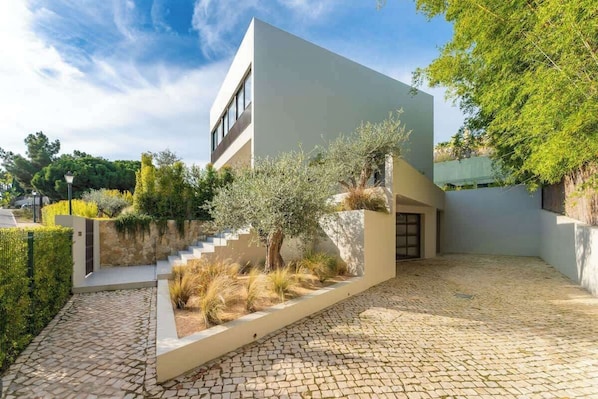Un edificio moderno con techo plano, un camino de piedra que conduce a la entrada y un pequeño jardín con un árbol y arbustos.