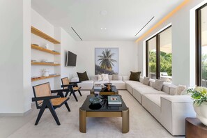 Un salon moderne avec un canapé, des fauteuils, une table basse et un téléviseur. On y trouve des étagères remplies de livres et d’objets décoratifs.