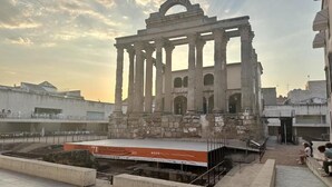 Ein antiker griechischer Tempel mit Säulen, einer Bühne und einem modernen Gebäude im Hintergrund.