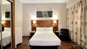 Ein Hotelzimmer mit einem großen Bett, einem Holzkopfteil, zwei Nachttischen mit Lampen, einem Spiegel und einem blumigen Vorhang.