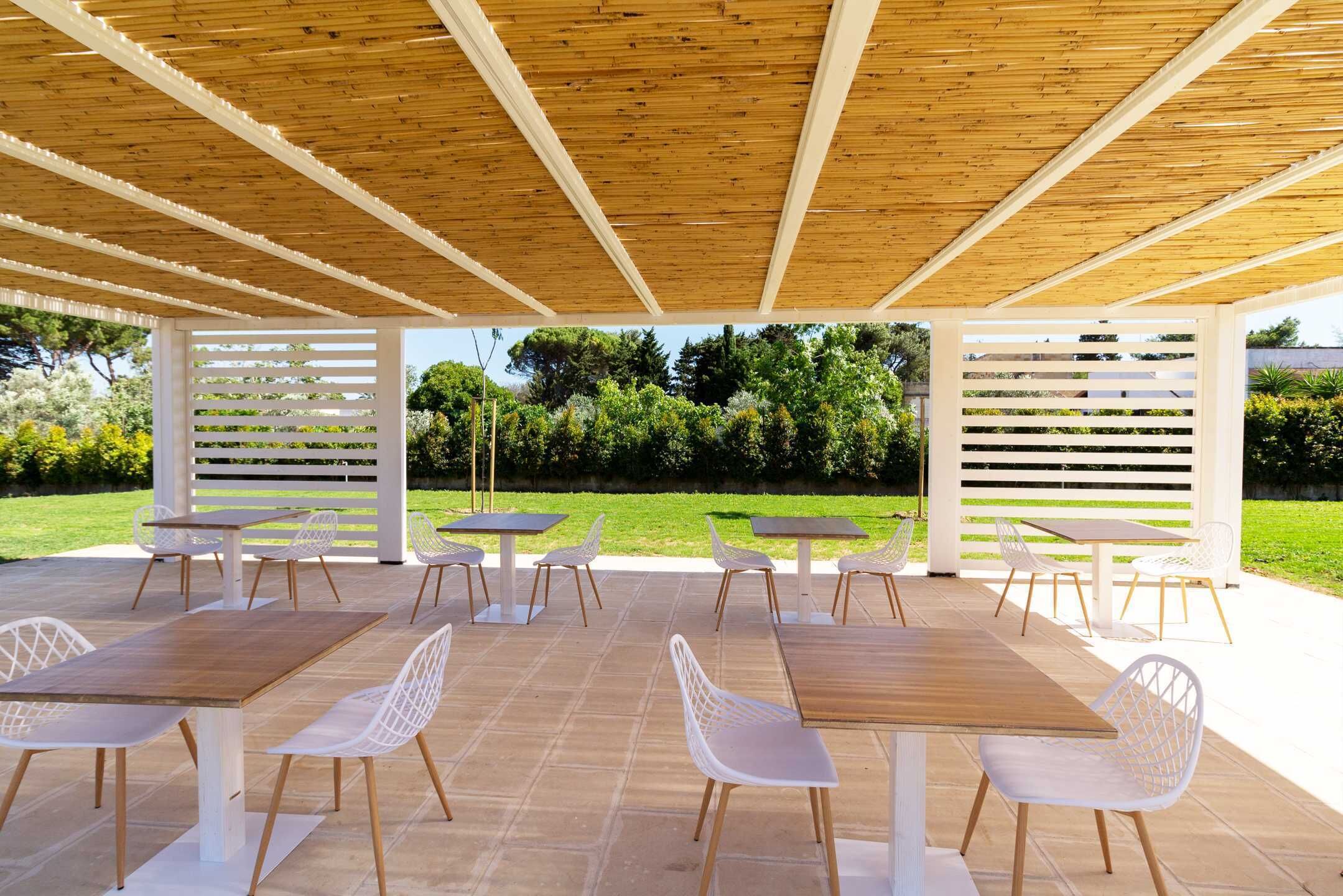 Une terrasse extérieure avec des tables en bois et des chaises blanches, entourée d’une pergola en bois et de panneaux de treillis blancs.
