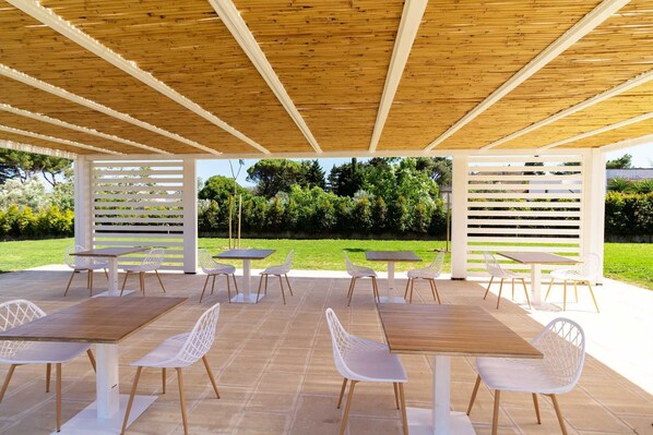 An outdoor patio with wooden tables and white chairs, surrounded by a wooden pergola and white lattice panels. - Sound Garden Charming Rooms (Melendugno)