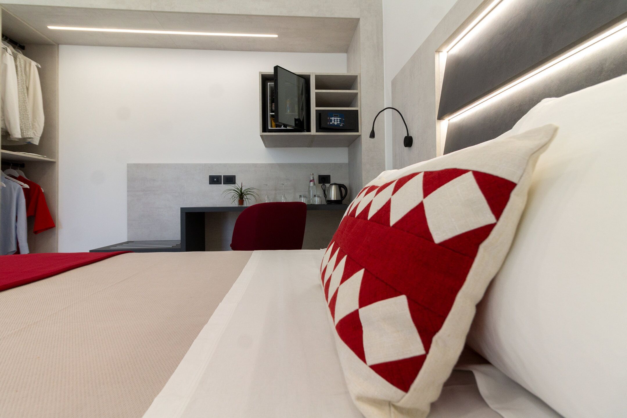 A modern hotel room with a bed, a red and white patterned pillow, a red chair, a small desk with a plant, and a built-in TV shelf.