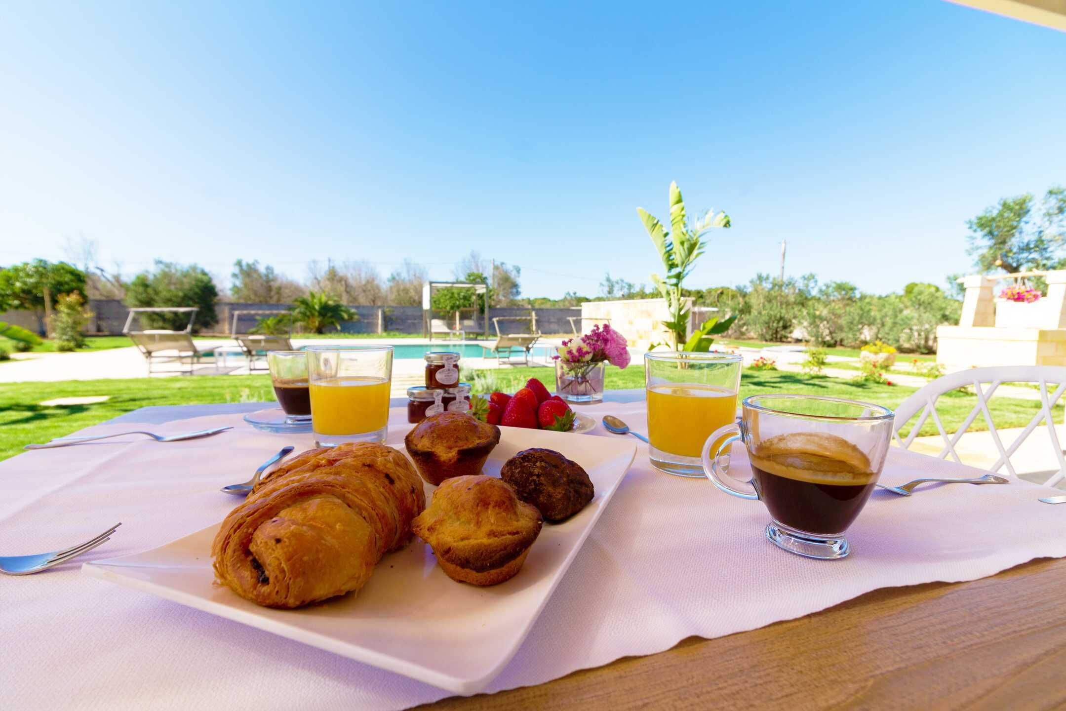A breakfast setting with croissants, muffins, and coffee on a table with a view of a pool and garden.
