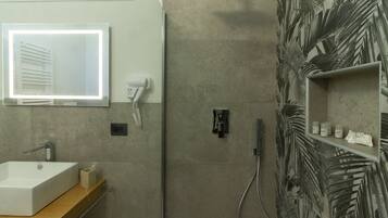 Un baño con un lavamanos rectangular, un espejo, un secador de pelo y una zona de ducha con diseño de hoja de palma en la pared.