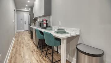 A narrow kitchen with a white countertop, teal bar stools, and a stainless steel trash can.