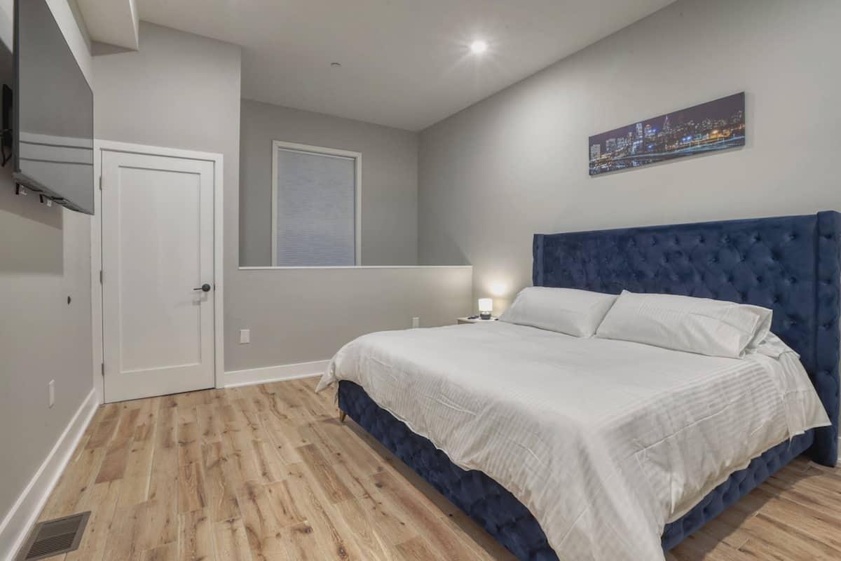 A bedroom with a large bed, a cityscape painting, and a window with blinds.