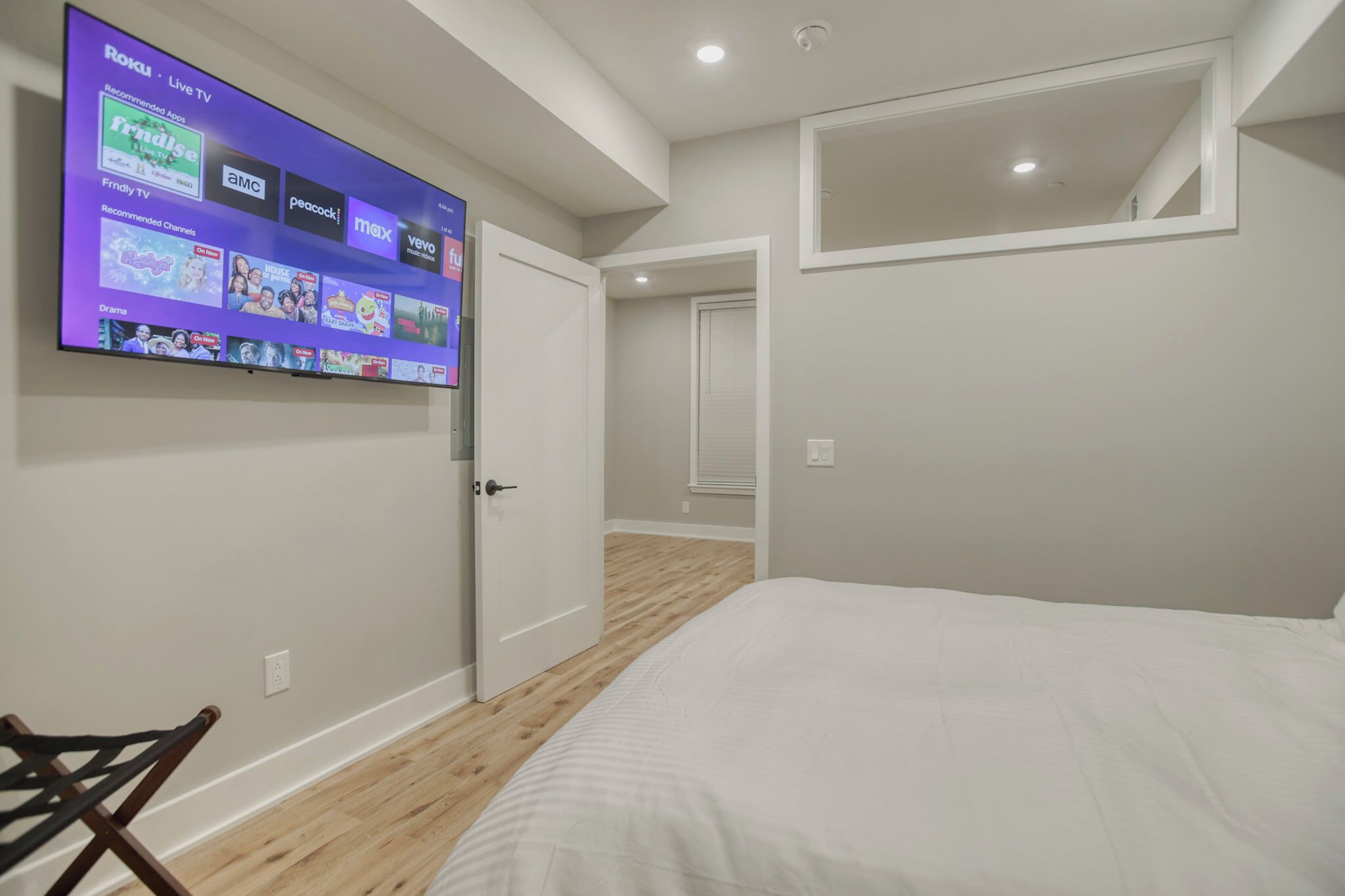 A bedroom with a bed, a TV mounted on the wall displaying various channels, and a door leading to another room.