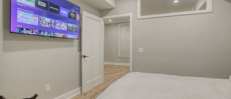 Un dormitorio con cama, televisor montado en la pared mostrando varios canales, y una puerta que da a otra habitación.