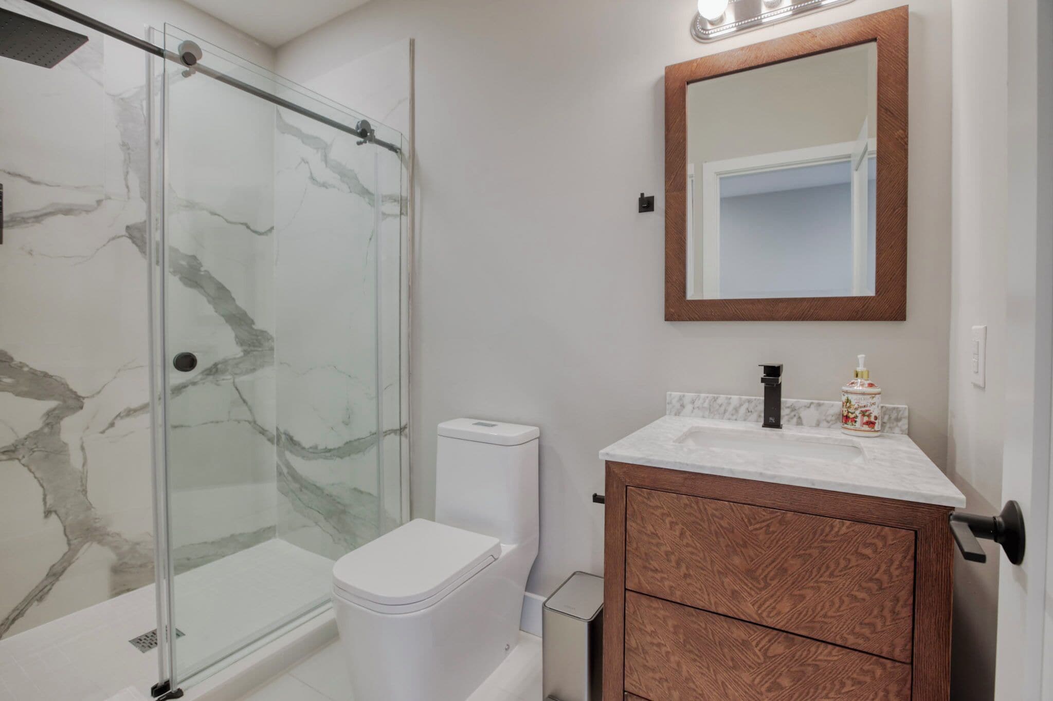 Un bagno con box doccia in vetro, un WC bianco, una cassettiera in legno con piano in marmo e uno specchio con cornice in legno.