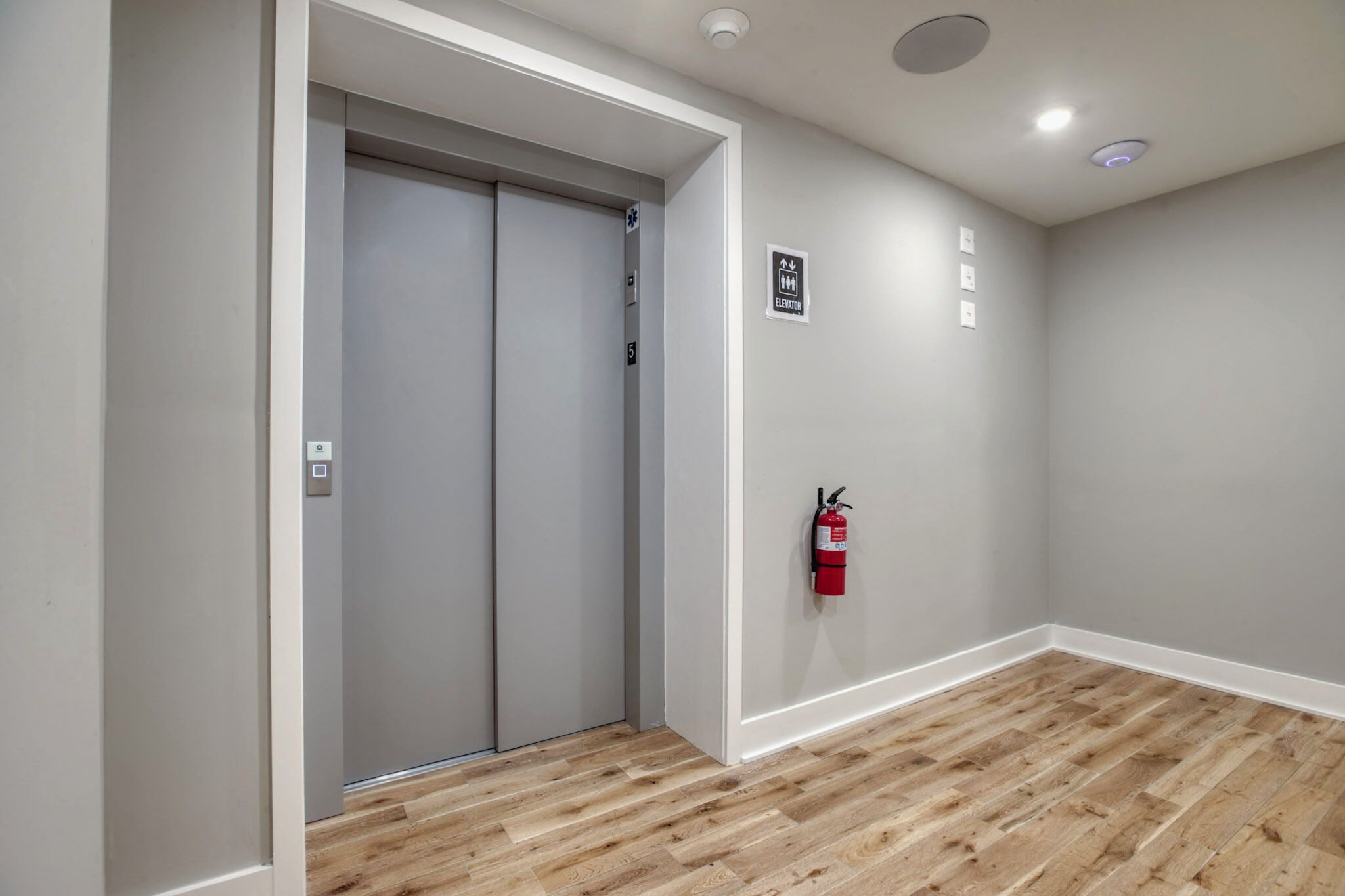 Un pasillo con piso de madera, un ascensor gris y un extintor en la pared.