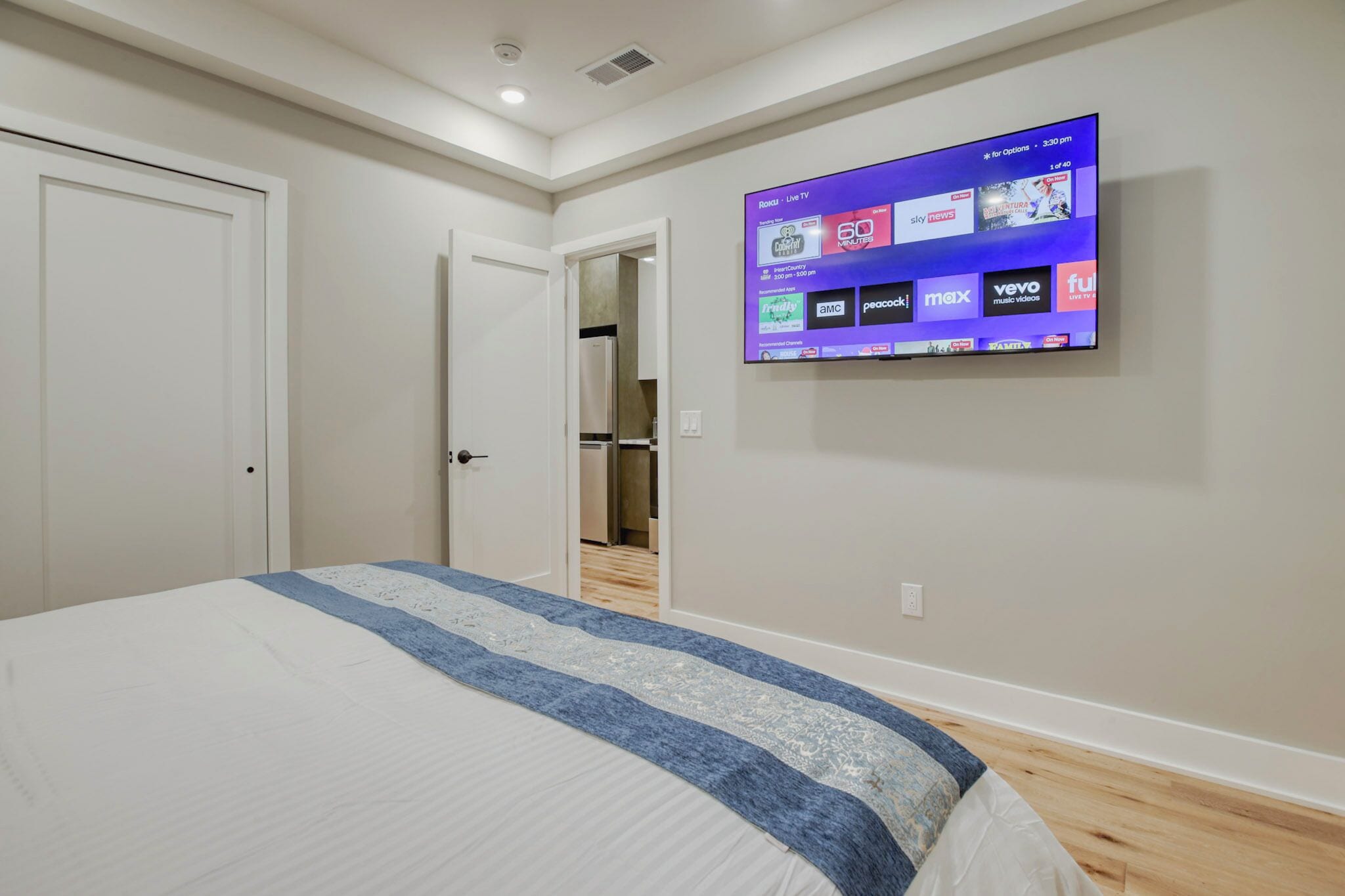 A bedroom with a large bed, a flat-screen TV mounted on the wall displaying various streaming services, and a door leading to a hallway.