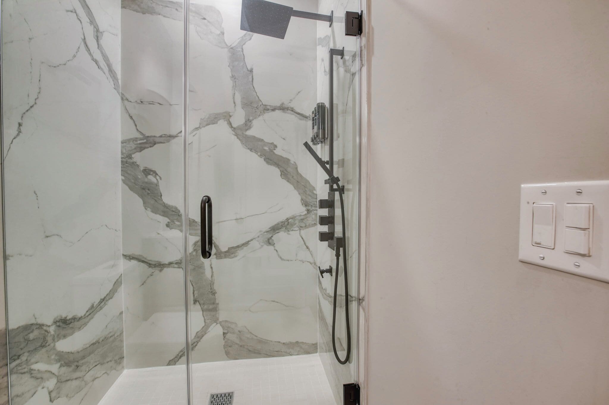 A glass-enclosed shower with a marble wall design, a handheld showerhead, and a wall-mounted control panel.