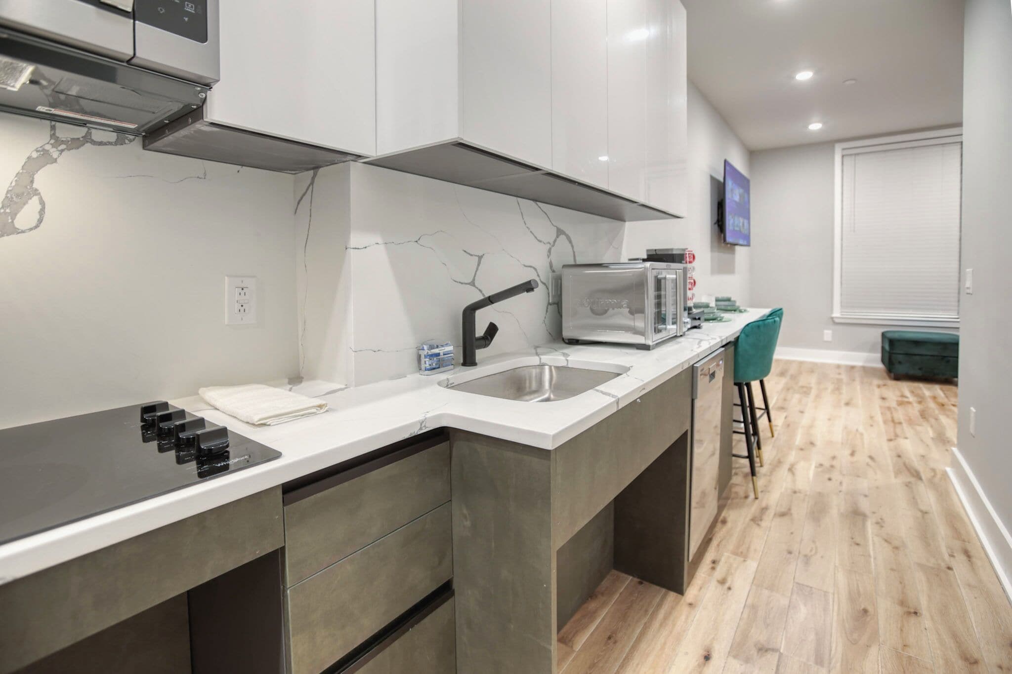 A modern kitchen with a built-in stove, a sink, and a microwave. There is a bar stool and a window with blinds in the adjacent dining area.