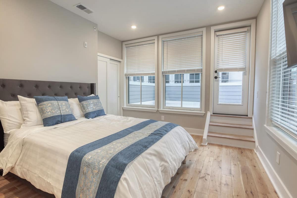 A bedroom with a large bed, a window with blinds, and a staircase leading to an upper level.