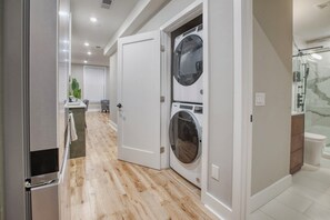 A laundry room with two stacked washing machines, a shower area, and a hallway leading to a dining area with a plant. - One Sky Loft (Philadelphia)