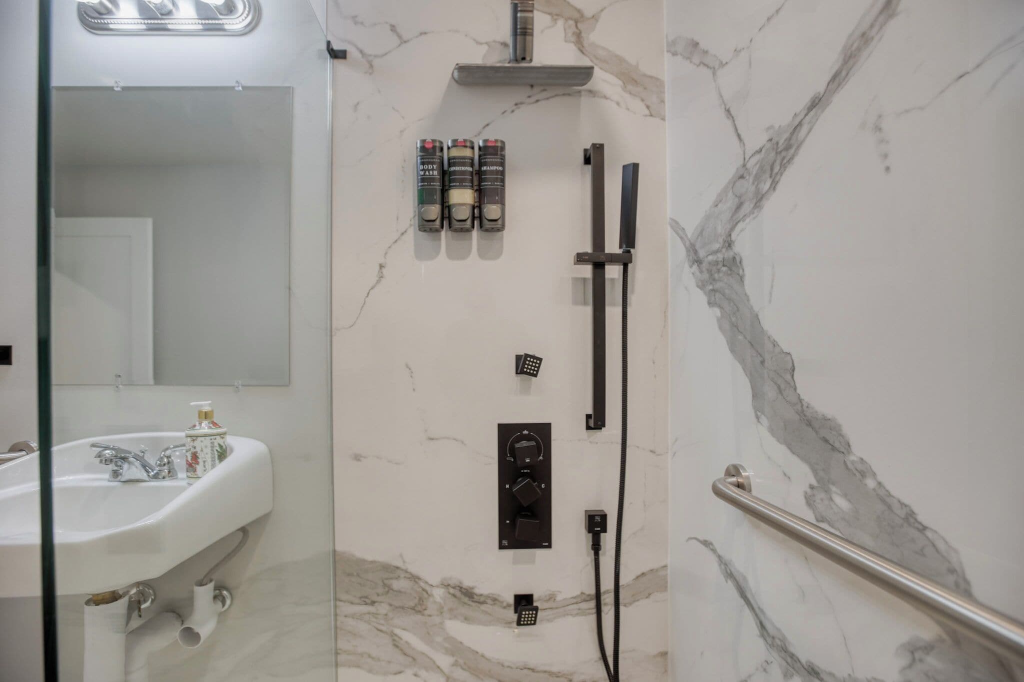 A bathroom with a marble wall, a sink, a mirror, and a shower with a handheld showerhead and shampoo bottles.