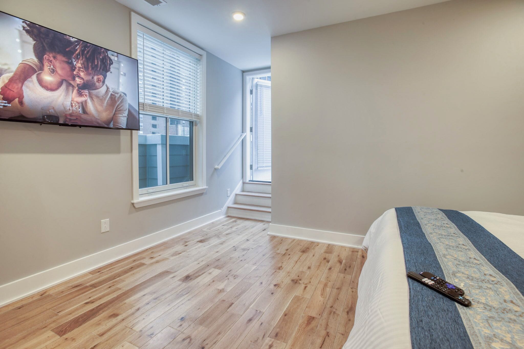 A bedroom with a large TV mounted on the wall, a wooden floor, a bed with a blue and white blanket, and a window with blinds.
