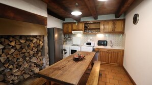 A rustic kitchen with a wooden table, a stone wall with firewood, and basic appliances. - Gîte ST Saturnin, Cézallier, Monts du Cantal, Santoire Valley (Saint-Saturnin)