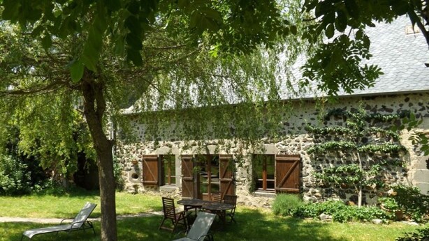 A stone building with a rustic exterior, a covered patio with a table and chairs, and a tree providing shade. - Gîte ST Saturnin, Cézallier, Monts du Cantal, Santoire Valley (Saint-Saturnin)