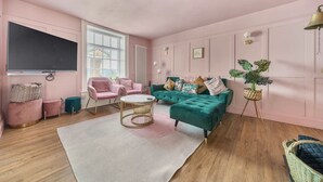 A living room with a large flat-screen TV, two pink armchairs, a teal sofa, a coffee table, and a potted plant. - Georgian Coach House in a Prime Location in Deal (Kent)