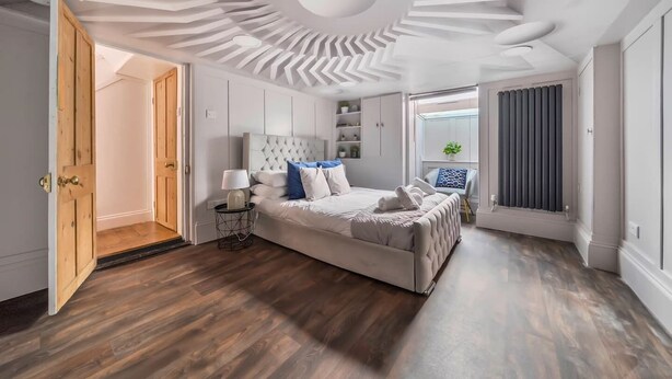 A modern bedroom with a tufted headboard, a bed with white bedding and blue accents, a bedside table with a lamp, and a window with blinds. - Georgian Coach House in a Prime Location in Deal (Kent)